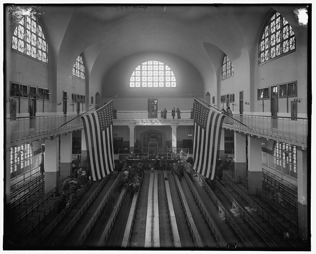 The early Ellis Island inspection room: long benches, railings, high windows and flags beneath the original plaster vault