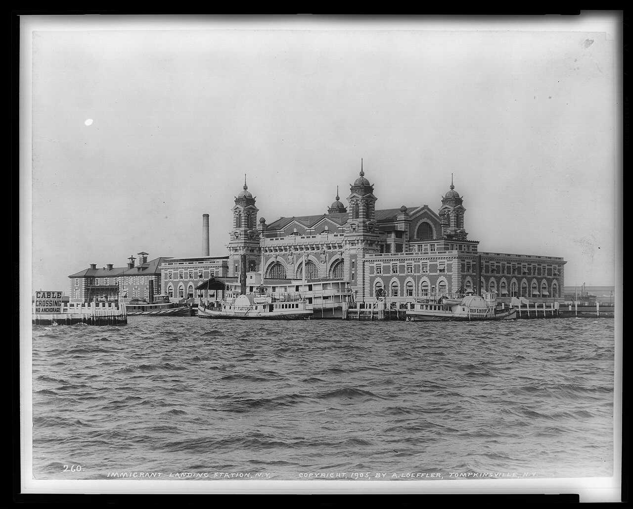 Ellis Island's main building viewed across water, with smaller vessels at its docks; copyright 1905, exact exposure date unknown